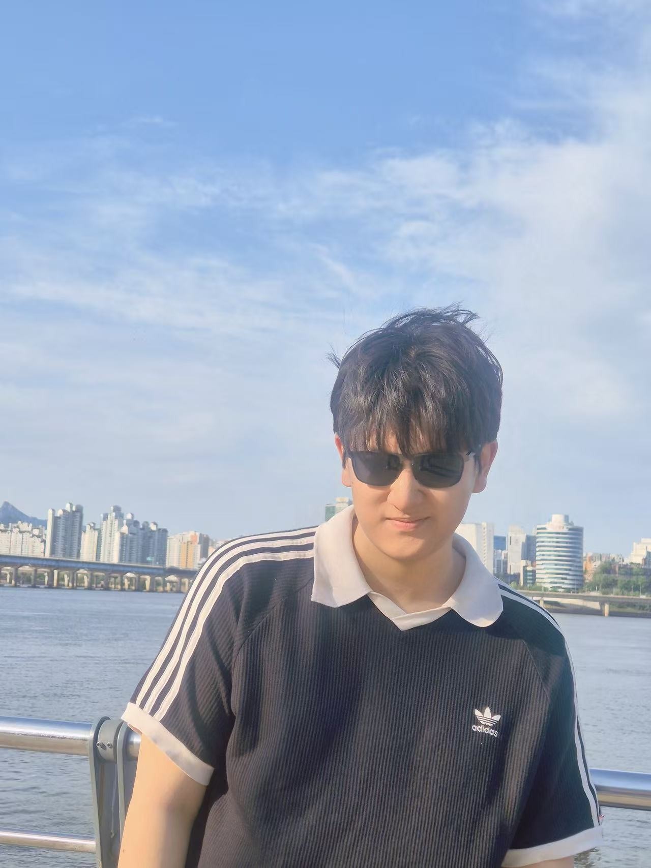 mioyoung戴着墨镜站在江边，身后是蓝天和城市